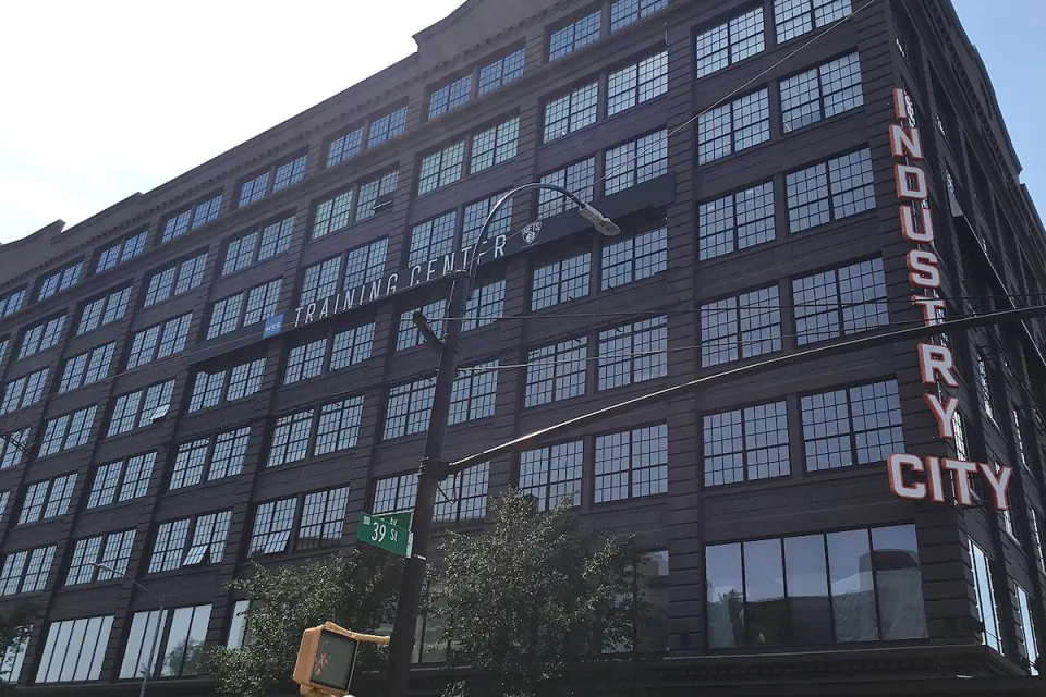 Looking up at black multi-story building with Industry City neon sign on corner.